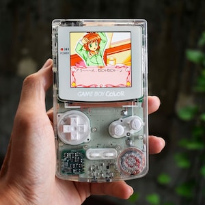 以下が含まれることがあります： 内部基板が見える透明なゲームボーイカラー携帯ゲーム機。白い十字キーと白いAボタン、Bボタンを備えています。画面にはアニメ調のキャラクターと日本語のテキストが表示されています。