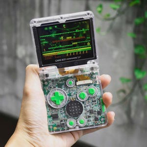 May include: A clear Game Boy Advance SP handheld console with a green and black screen displaying a retro video game. The console's internal circuit board is visible. Green buttons and a green directional pad are featured on the console.