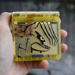 May include: A transparent yellow Game Boy Advance SP case with a black graphic of a monster's face. The case has a Nintendo logo and internal components are visible. The device is held in a hand.