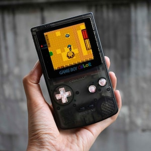 Puede incluir: Una consola de juegos portátil Game Boy Color negra con una carcasa translúcida. La pantalla muestra un videojuego pixelado con un fondo amarillo y un personaje en el centro. La consola tiene un pad direccional en forma de cruz blanco y dos botones rosas.