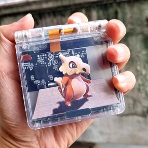 Könnte beinhalten: Eine durchsichtige Game Boy Color Handheld-Spielkonsole aus Plastik mit einem Bildschirm, der ein Cartoon-Bild eines braunen und weißen Pokemon-Charakters zeigt. Der Charakter steht auf einer hellbraunen Oberfläche. Der Text "L-R TRIGGER" ist an der Seite der Konsole sichtbar.
