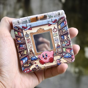 May include: A square, clear plastic device with a colorful illustration of a room with toys and a pink cartoon character. The design includes bookshelves, airplanes, and toy cars. The device is held in a hand.