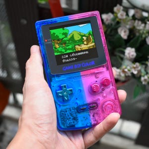 Puede incluir: Una consola de videojuegos portátil Game Boy Color con una carcasa transparente de dos tonos, con un lado izquierdo azul y un lado derecho rosa. La pantalla muestra un paisaje pixelado colorido con texto en japonés y los botones a juego.