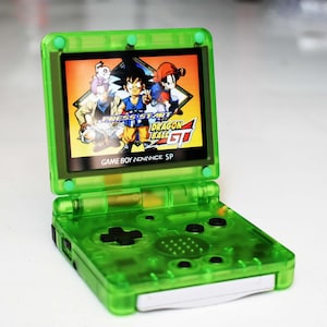 May include: A transparent green Game Boy Advance SP handheld console, open to display a colorful Dragon Ball GT game screen. The console features a directional pad, buttons, and the text "GAME BOY ADVANCE SP".