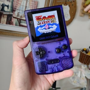 Puede incluir: Una consola de juegos portátil Game Boy Color de color morado translúcido con una cruceta negra, dos botones de acción negros etiquetados como A y B, y botones Select y Start negros. La pantalla muestra el título Zelda no Densetsu Yume wo Miru Shima DX.