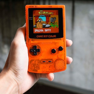 Puede incluir: Una consola de videojuegos portátil Game Boy Color naranja con una pantalla en blanco y negro. El juego "The Flintstones: Special Duty!" se muestra en la pantalla.