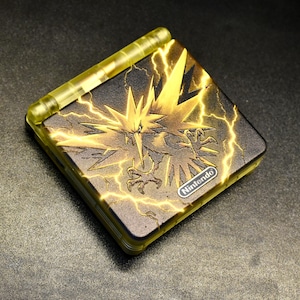 Può includere: Un dispositivo di gioco portatile con un design nero e oro. La parte superiore presenta un'illustrazione dorata di un uccello con fulmini e il logo Nintendo. Il dispositivo ha una cerniera e una cornice gialle.