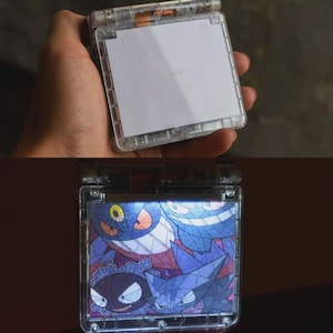 May include: A transparent, handheld device with a hinged top, showing a white screen. The device is illuminated, revealing a colourful graphic of cartoon characters in purple, blue, and yellow. The device is held in a person's hand.