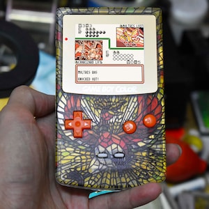 以下が含まれることがあります： 赤、オレンジ、黄色の鳥が描かれたステンドグラスのデザインのゲームボーイカラー携帯ゲーム機。画面には「MOLTRES HAS KNOCKED OUT!」というテキストが表示されたポケモンバトルが表示されています。