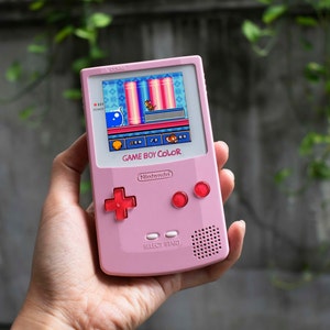 May include: A pink Game Boy Color handheld video game console with a game displayed on the screen. The console has red buttons and a directional pad. The screen displays a colorful 2D game scene.