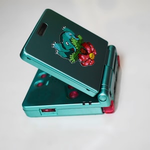 May include: Teal handheld games console with a hinged design. The top cover has a sticker of a cartoon character with a flower. Red buttons and a white surface complete the design.