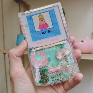Op de afbeelding: Een transparante Game Boy Color handheld spelcomputer met een lichtgroene interne behuizing en roze knoppen. Het scherm toont een korrelige afbeelding van Prinses Peach met het cijfer 7 en een 0/9 teller. De tekst GAME BOY COLOR is in het roze gedrukt.