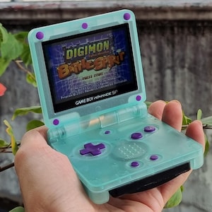 Op de afbeelding: Een turquoise Game Boy Advance SP handheld console met paarse knoppen en accenten. Het scherm toont het spel Digimon Battle Spirit. De console wordt vastgehouden voor een achtergrond van groen.