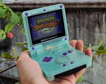 Könnte beinhalten: Türkisfarbene Game Boy Advance SP-Konsole mit lila Knöpfen und Akzenten. Der Bildschirm zeigt das Spiel Digimon Battle Spirit. Die Konsole wird vor einem Hintergrund aus grünem Laub gehalten.