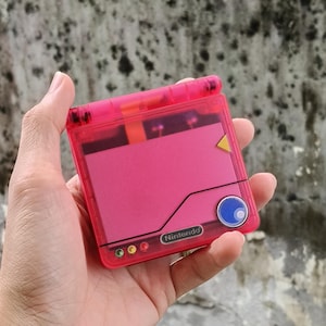 May include: A translucent hot pink handheld gaming console with a clamshell design. The device features a solid pink rectangular panel on the lid, a small yellow triangle accent, a blue circular button, and the Nintendo logo printed in silver.
