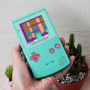 Puede incluir: Una consola de juegos portátil Game Boy Color de color verde azulado con botones rosas. La pantalla muestra un videojuego pixelado con un fondo azul y obstáculos morados y marrones. El texto "GAME BOY COLOR" es visible en la consola.