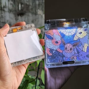 Könnte beinhalten: Transparente Kunststoff-Handheld-Spielhülle. Auf der linken Seite befindet sich ein weißer Einsatz, während die rechte Seite eine Grafik im Buntglasstil mit Cartoon-Charakteren in Rosa, Blau und Lila zeigt. Das Wort "tendo" ist sichtbar.