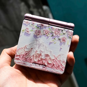 Könnte beinhalten: Ein tragbares Spielgerät mit floralem und bergigem Design. Das Gerät hat eine hellviolette Farbe mit einem Klappdesign. Die Vorderseite zeigt eine Berglandschaft mit rosa und weißen Blumen.