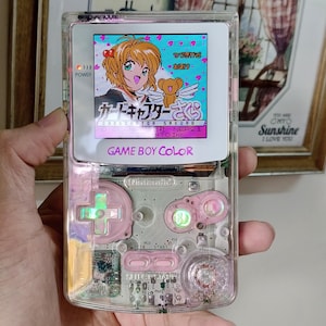 May include: A transparent Game Boy Color handheld gaming console featuring a clear shell, light pink buttons, and a directional pad. The screen displays a colorful anime-style graphic with Japanese text and the title Cardcaptor Sakura.