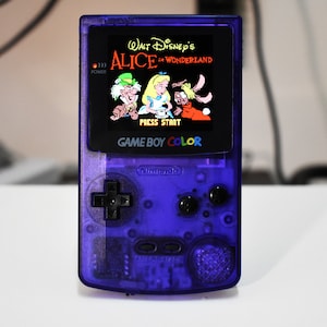 Puede incluir: Una consola de videojuegos portátil Game Boy Color de color morado con una pantalla que muestra el juego "Walt Disney's Alice in Wonderland". El juego presenta a Alice, el Sombrerero Loco y el Conejo Blanco.