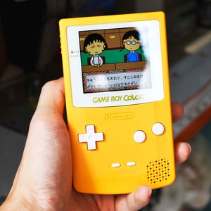 Puede incluir: Una consola de videojuegos portátil Game Boy Color amarilla con una placa frontal blanca. La pantalla muestra un juego con dos personajes y texto japonés. El texto en la consola dice "GAME BOY COLOR" y "Nintendo".