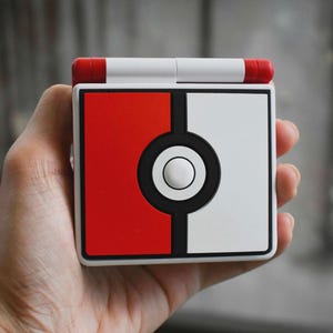 May include: A handheld gaming device in red and white, with a black circle design in the centre. The device has a white top with red accents and is held in a hand.