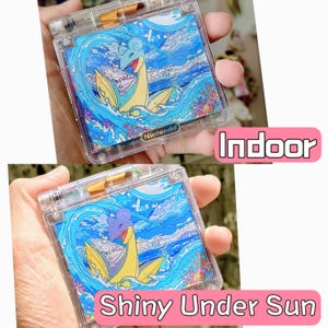 Könnte beinhalten: Eine durchsichtige, transparente Gehäusehülle für eine tragbare Spielkonsole mit einer lebendigen Grafik eines blauen und gelben Lapras-Wesens, das in Meereswellen mit Korallenriffen schwimmt. Das Nintendo-Logo ist am unteren Rand der Konsole sichtbar.