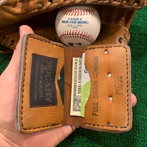 May include: A brown leather wallet with the text "MOCABY" and "FULL GRAIN COWHIDE" is open, displaying a credit card and a dollar bill. A baseball and a baseball glove are in the background.