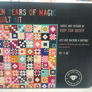 Peut inclure: Une boîte noire de kit de courtepointe avec le texte "TEN YEARS OF MAGIC QUILT KIT". Le motif de la courtepointe présente un motif patchwork coloré avec diverses formes et couleurs. La boîte indique également "FABRIC AND PATTERN BY RUBY STAR SOCIETY" et la taille finale de la courtepointe est de 203 cm x 203 cm.