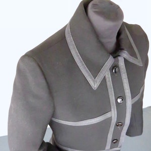 Può includere: Una giacca nera a maniche lunghe con colletto e chiusura con bottoni. La giacca ha una bordatura bianca sui bordi e sul davanti.