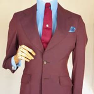 Pode incluir: Um blazer bordô com um fecho de botão único e um bolso de cada lado. O blazer é usado sobre uma camisa azul claro com uma gravata vermelha.