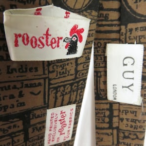 Może przedstawiać: Brązowo-czarny krawat z tkaniny z wzorem koguta i napisem "rooster" nadrukowanym na białej metce. Krawat ma również białą metkę z nadrukowanym tekstem "GUY LONDON".