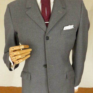 Pode incluir: Casaco de fato cinzento com camisa branca e gravata bordô. O casaco tem fecho de dois botões, lenço de bolso e lapela entalhada. O fato é exibido num manequim de madeira.