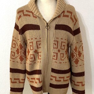 May include: A beige knit zip-up cardigan featuring a shawl collar and a repeating geometric pattern in dark brown and rust orange. The sweater has long sleeves, a full-length metal zipper with a circular pull, and a thick, ribbed texture throughout.
