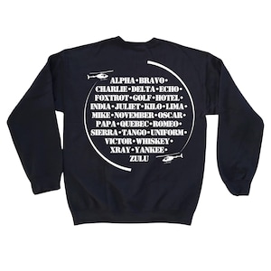 Könnte beinhalten: Ein marineblaues Sweatshirt mit einem weißen Kreis-Motiv auf der Rückseite. Das Kreis-Motiv zeigt das NATO-phonetische Alphabet in weißer Schrift, mit einem Hubschrauber-Motiv oben und unten im Kreis.