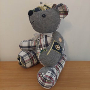 Pu&ograve; includere: Un orsacchiotto di peluche grigio e bianco a patchwork con un nastro bianco intorno al collo. L'orso ha un bottone sul petto ed &egrave; fatto di diversi tessuti.