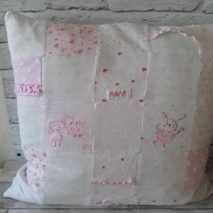 May include: A square patchwork cushion in white and pink. It features embroidered images of a bunny and a bear, with the text "Pretty Little Angel". The cushion is made of various fabric squares, creating a patchwork design.