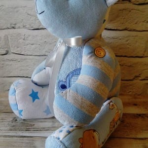 Pu&ograve; includere: Un orsacchiotto a strisce blu e bianche con un nastro bianco intorno al collo. L'orso &egrave; fatto di vestiti per bambini riciclati e ha un motivo a stelle sulle gambe. L'orso &egrave; seduto su una superficie di legno.
