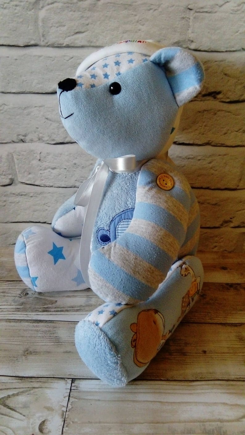 Pu&ograve; includere: Un orsacchiotto a strisce blu e bianche con un nastro bianco intorno al collo. L'orso &egrave; fatto di vestiti per bambini riciclati e ha un motivo a stelle sulle gambe. L'orso &egrave; seduto su una superficie di legno.