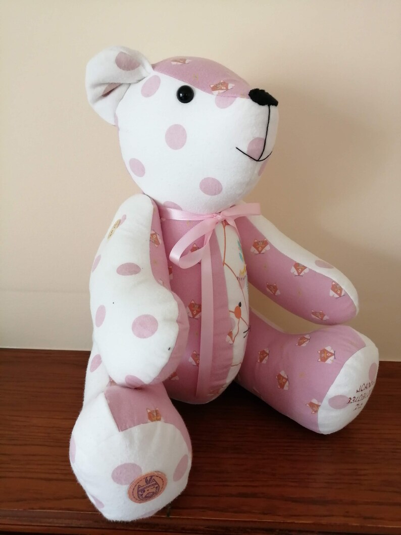 Pu&ograve; includere: Un orsacchiotto rosa e bianco a pois con un nastro rosa intorno al collo. L'orsacchiotto &egrave; seduto su una superficie di legno.