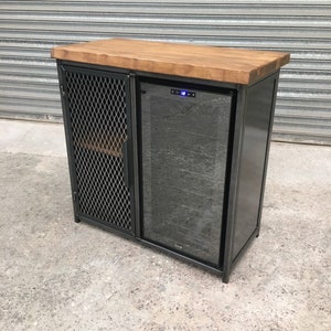 Op de afbeelding: Een zwarte metalen kast met een houten blad en een ingebouwde wijnkoeler. De kast heeft een draadgaasdeur aan de linkerkant en een glazen deur aan de rechterkant. De wijnkoeler heeft een digitaal display en een glazen deur.