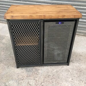 Op de afbeelding: Een zwarte metalen kast met een houten blad en een ingebouwde wijnkoeler. De kast heeft een draadgaasdeur aan de linkerkant en een glazen deur aan de rechterkant. De wijnkoeler heeft een digitaal temperatuurdisplay dat "12" aangeeft.