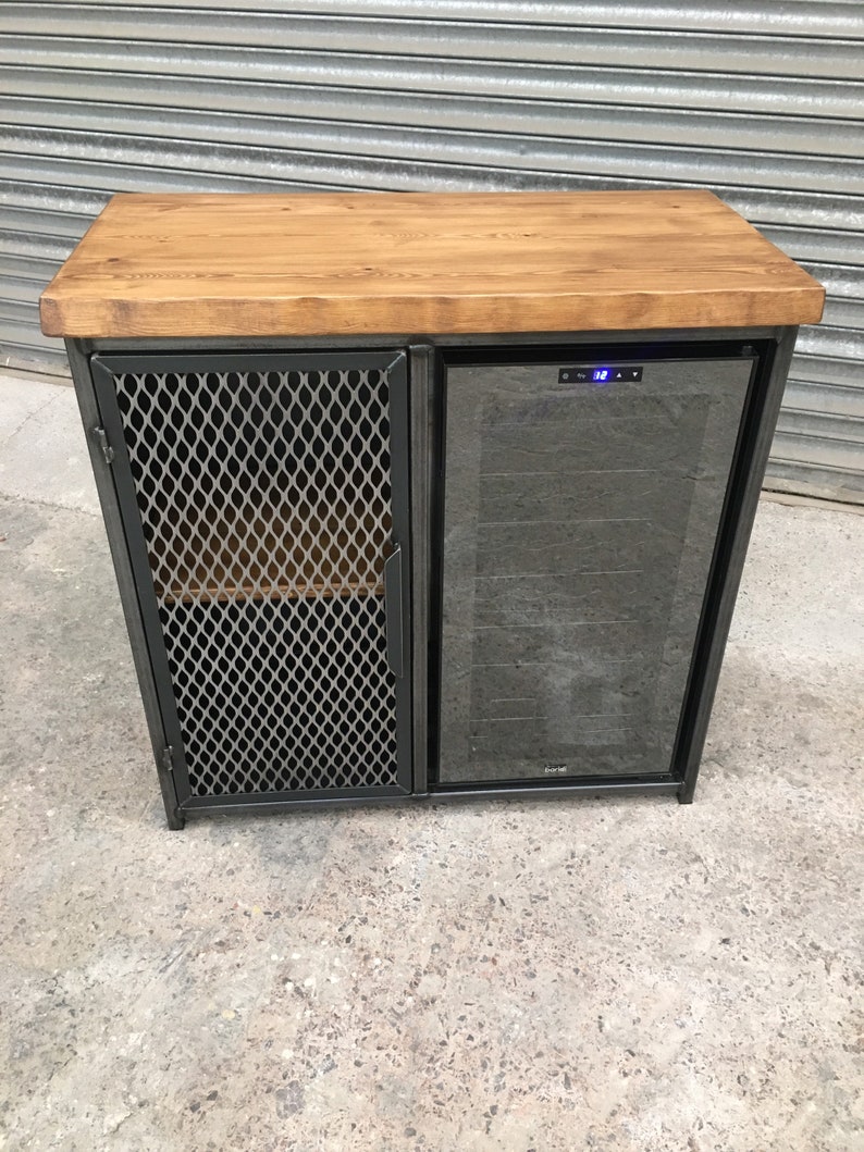 Op de afbeelding: Een zwarte metalen kast met een houten blad en een ingebouwde wijnkoeler. De kast heeft een draadgaasdeur aan de linkerkant en een glazen deur aan de rechterkant. De wijnkoeler heeft een digitaal temperatuurdisplay dat "12" aangeeft.