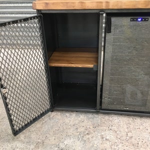 Op de afbeelding: Een zwarte metalen kast met een houten bovenblad en een glazen deur. De kast heeft een draadgaasdeur en een plank binnenin. De kast staat op een betonnen vloer.