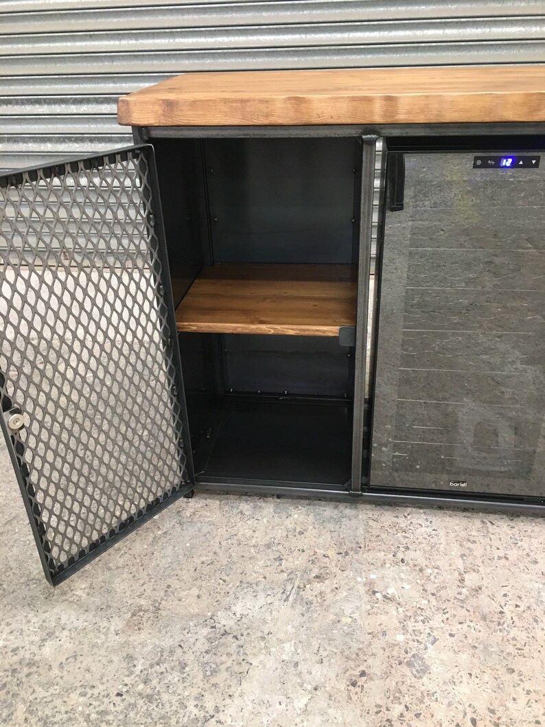 Op de afbeelding: Een zwarte metalen kast met een houten bovenblad en een glazen deur. De kast heeft een draadgaasdeur en een plank binnenin. De kast staat op een betonnen vloer.