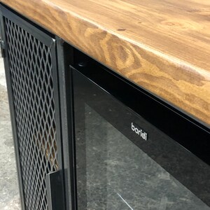 Pode incluir: Um m&oacute;vel com tampo de madeira natural de veio vis&iacute;vel e estrutura de metal preto. A unidade inclui uma porta lateral com um painel de malha met&aacute;lica em padr&atilde;o de diamante e uma sec&ccedil;&atilde;o de vidro com a marca baridi em letras brancas.