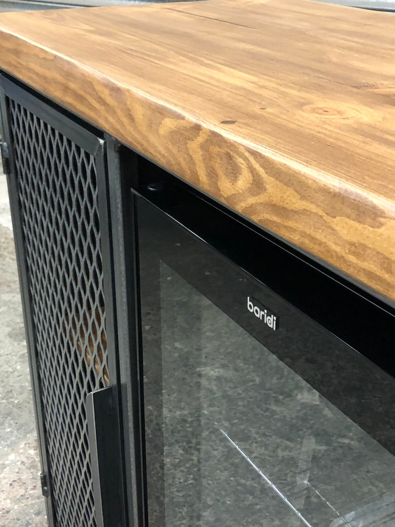 Pode incluir: Um m&oacute;vel com tampo de madeira natural de veio vis&iacute;vel e estrutura de metal preto. A unidade inclui uma porta lateral com um painel de malha met&aacute;lica em padr&atilde;o de diamante e uma sec&ccedil;&atilde;o de vidro com a marca baridi em letras brancas.