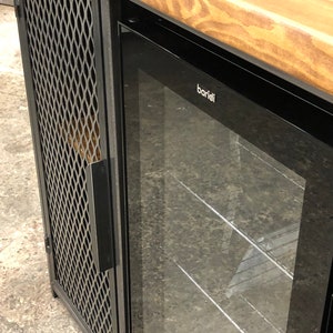 Pode incluir: Um m&oacute;vel com tampo de madeira natural, estrutura de metal preto e uma porta lateral com painel de malha met&aacute;lica em padr&atilde;o de losango. Ao lado da porta de malha, h&aacute; um refrigerador de bebidas com porta de vidro e a marca baridi no topo.