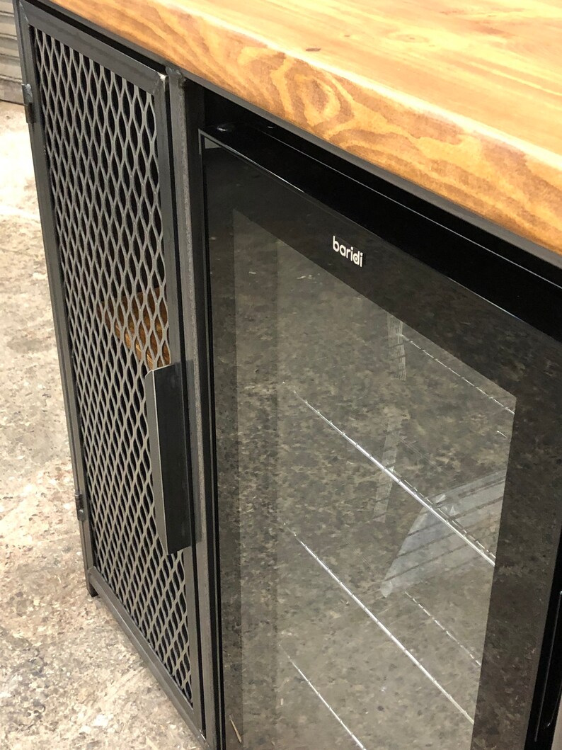 Pode incluir: Um m&oacute;vel com tampo de madeira natural, estrutura de metal preto e uma porta lateral com painel de malha met&aacute;lica em padr&atilde;o de losango. Ao lado da porta de malha, h&aacute; um refrigerador de bebidas com porta de vidro e a marca baridi no topo.