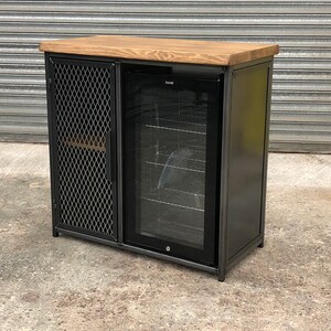 Pode incluir: Um m&oacute;vel retangular com estrutura de metal escuro, tampo de madeira natural e porta de malha met&aacute;lica preta &agrave; esquerda. O lado direito cont&eacute;m um refrigerador de bebidas embutido com porta de vidro, prateleiras de arame e acabamento preto.